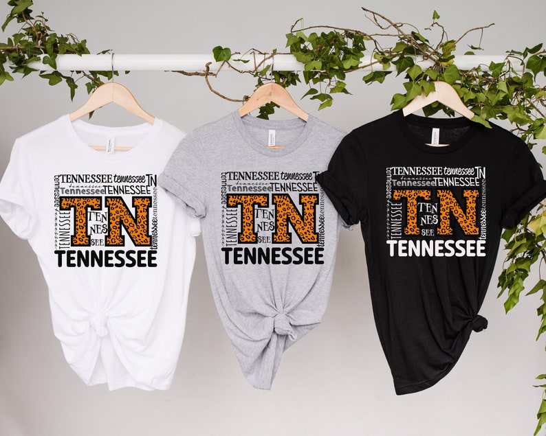 May include: Three t-shirts with a graphic design featuring the letters "TN" in a large font with a leopard print pattern. The letters are surrounded by the word "Tennessee" repeated multiple times in a smaller font. The t-shirts are white, grey, and black.