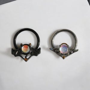 Puede incluir: Dos piercings para la nariz. Uno es negro con un dije en forma de murciélago que sostiene una gema colorida. El otro es plateado con un dije decorativo que sostiene una gema con tonos rosas y azules. Ambos anillos tienen un diseño circular.