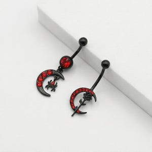 Puede incluir: Dos piercings negros para el ombligo. Uno presenta una media luna con un dije de araña y gemas rojas. El otro tiene una media luna con una rosa y gemas rojas. Ambos tienen un diseño de barra curva con una bola negra en la parte superior.