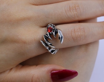 May include: A silver ring with a claw design, featuring a red gemstone. The ring has sharp, pointed details and is worn on a finger. The gemstone is a vibrant red colour.