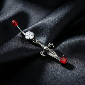 May include: A silver-colored belly button ring with a sword design. The curved barbell features a red bead at the top and a clear, round gem in the center. The sword has a skull and heart-shaped red gem at the bottom.