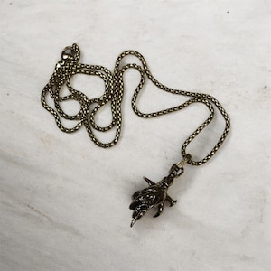 Könnte beinhalten: Eine bronzefarbene Halskette mit einem detaillierten Handanhänger. Die Kette hat ein strukturiertes, gewebtes Aussehen. Der Anhänger ist eine detaillierte Hand mit scharfen Krallen. Die Halskette wird auf einer weiß-grauen Marmoroberfläche präsentiert.