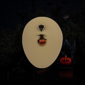 Puede incluir: Anillos para el ombligo con temática de Halloween. Un anillo presenta un pequeño murciélago negro con ojos rojos, y el otro es una calabaza sonriente. Una pequeña calabaza naranja con una cara tallada se encuentra en el fondo. El fondo es una escena de bosque oscuro.