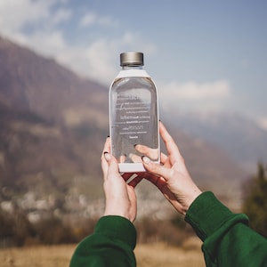 Könnte beinhalten: Eine klare Glaswasserflasche mit weißem Verschluss und einem Etikett mit der Aufschrift "leben kraft erfrichend" und "freude".