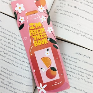 Pode incluir: Marcador de livros rosa com um design de garrafa de sumo e o texto "I'm Peelin' This Book". O marcador apresenta fatias de laranja, pêssegos e flores brancas. Está colocado num livro aberto.