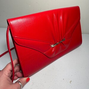 Peut inclure: Un sac pochette rectangulaire rouge vif avec une surface texturée et une finition brillante. Le rabat avant présente un détail plissé central maintenu par une fine barre métallique dorée. Une fine bandoulière rouge assortie est fixée sur le côté.
