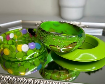 May include: Three bangle bracelets. One is clear with embedded multicolored circular confetti. Another is bright green with gold leaf flakes and black line art of butterflies. The third is a solid, smooth, bright lime green bangle.