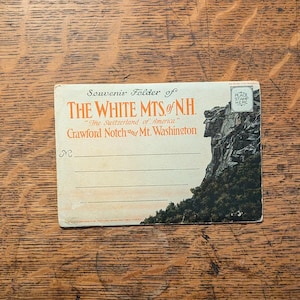 May include: A vintage postcard featuring a scenic view of the White Mountains of New Hampshire, with the text "Souvenir Folder of THE WHITE MTS. of NH. "The Switzerland of America" Crawford Notch and Mt. Washington".