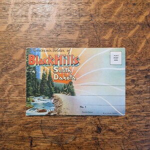 May include: Vintage postcard featuring a scenic view of the Black Hills in South Dakota. The postcard has a colorful design with text that reads "Souvenir Folder of Black Hills South Dakota." Includes a place for a stamp.