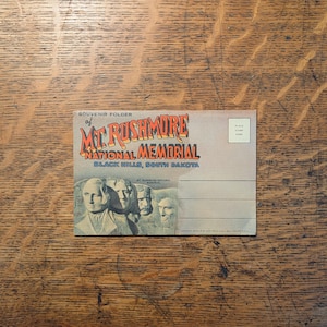 May include: A vintage souvenir postcard featuring Mount Rushmore National Memorial in the Black Hills of South Dakota. The postcard has a faded color scheme and includes the text "Souvenir Folder of Mt. Rushmore National Memorial".