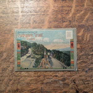 May include: Vintage postcard titled "Souvenir Folder of Mohawk Trail, Mass." featuring a scenic view of a road winding through a forested area. The postcard includes an illustration of a Native American figure and decorative borders.