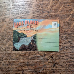May include: Vintage postcard featuring a scenic view of Lake Placid, N.Y., with a waterfall and lush greenery. The text reads "Lake Placid N.Y. Premier Resort in the Adirondack Mts." The postcard has a place for a stamp and address.