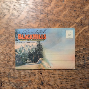 May include: Vintage postcard featuring a scenic view of the Black Hills in South Dakota. The postcard has the words "Beautiful Black Hills" in orange and blue, with a landscape image of a road and mountains. The postcard is from 1931.