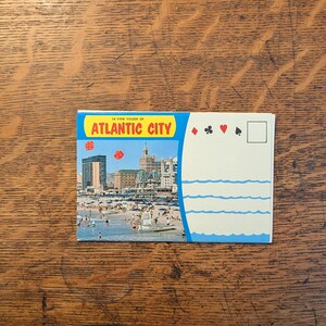 May include: A vintage postcard featuring a colorful image of Atlantic City, with the text "14 View Folder of Atlantic City" in yellow and blue. The postcard shows a beach scene with buildings and people. The right side has playing card symbols.