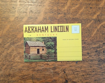 May include: A vintage postcard with the text "ABRAHAM LINCOLN IN KENTUCKY" in yellow letters. The postcard features a small wooden cabin and a split-rail fence. The words "VIEWS IN NATURAL COLOR" are also printed on the postcard.