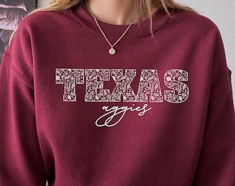 May include: A maroon sweatshirt with a white design that says "TEXAS Aggies" in a decorative font. The sweatshirt is being worn by a person with a plaid skirt.