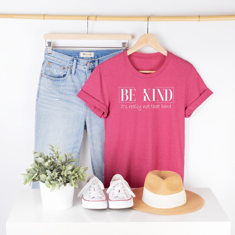 May include: A pink t-shirt with the text "BE KIND it's really not that hard" in white. The shirt is on a hanger with a pair of blue jeans, a plant, and a straw hat.