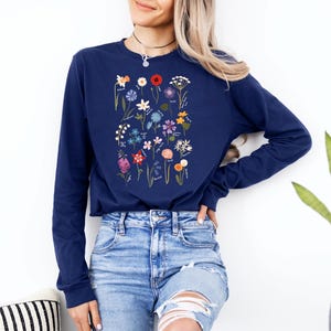 May include: A navy blue long-sleeved shirt with a floral print featuring a variety of flowers and their names. The shirt is being worn by a person with long blonde hair and blue jeans.