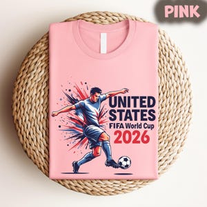 Pode incluir: Camiseta rosa claro com um jogador de futebol e o texto "UNITED STATES FIFA World Cup 2026" em vermelho e branco. A camiseta est&aacute; dobrada sobre uma superf&iacute;cie redonda de vime.