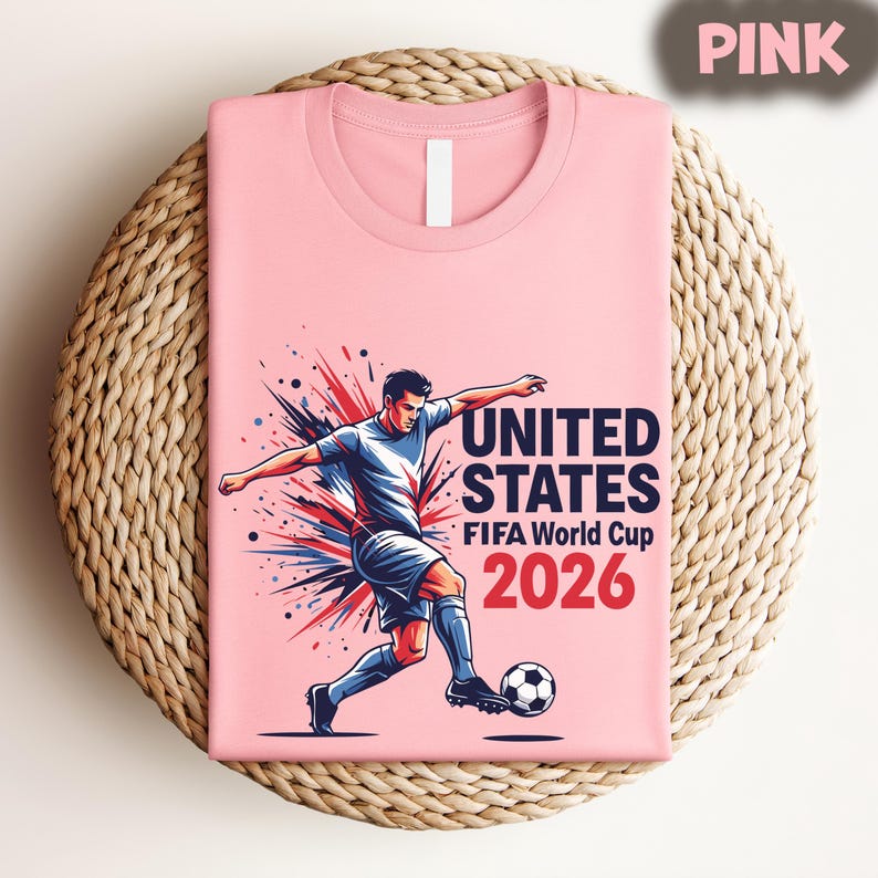Pode incluir: Camiseta rosa claro com um jogador de futebol e o texto "UNITED STATES FIFA World Cup 2026" em vermelho e branco. A camiseta est&aacute; dobrada sobre uma superf&iacute;cie redonda de vime.