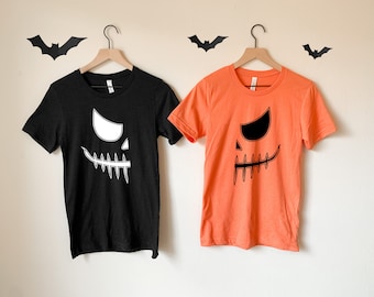 May include: Two t-shirts featuring a jack-o'-lantern face design. One shirt is black with a white jack-o'-lantern face and the other is orange with a black jack-o'-lantern face.