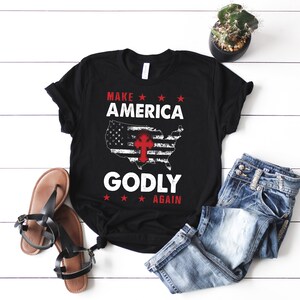 May include: Black t-shirt with a white and red American flag design and the text "Make America Godly Again".