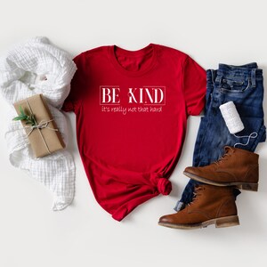 May include: Red t-shirt with white text that reads "BE KIND it's really not that hard". The shirt is laid flat on a white surface with a white scarf, a brown gift box, a pair of blue jeans, and brown boots.