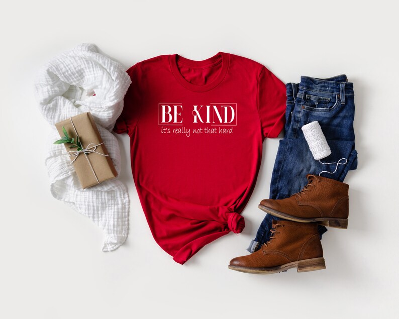 May include: Red t-shirt with white text that reads "BE KIND it's really not that hard". The shirt is laid flat on a white surface with a white scarf, a brown gift box, a pair of blue jeans, and brown boots.
