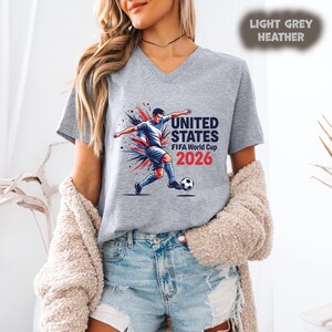 Pode incluir: T-shirt cinza claro mesclado com decote em V com um gr&aacute;fico de um jogador de futebol chutando uma bola, com o texto "UNITED STATES FIFA World Cup 2026". O design inclui detalhes em vermelho, branco e azul.