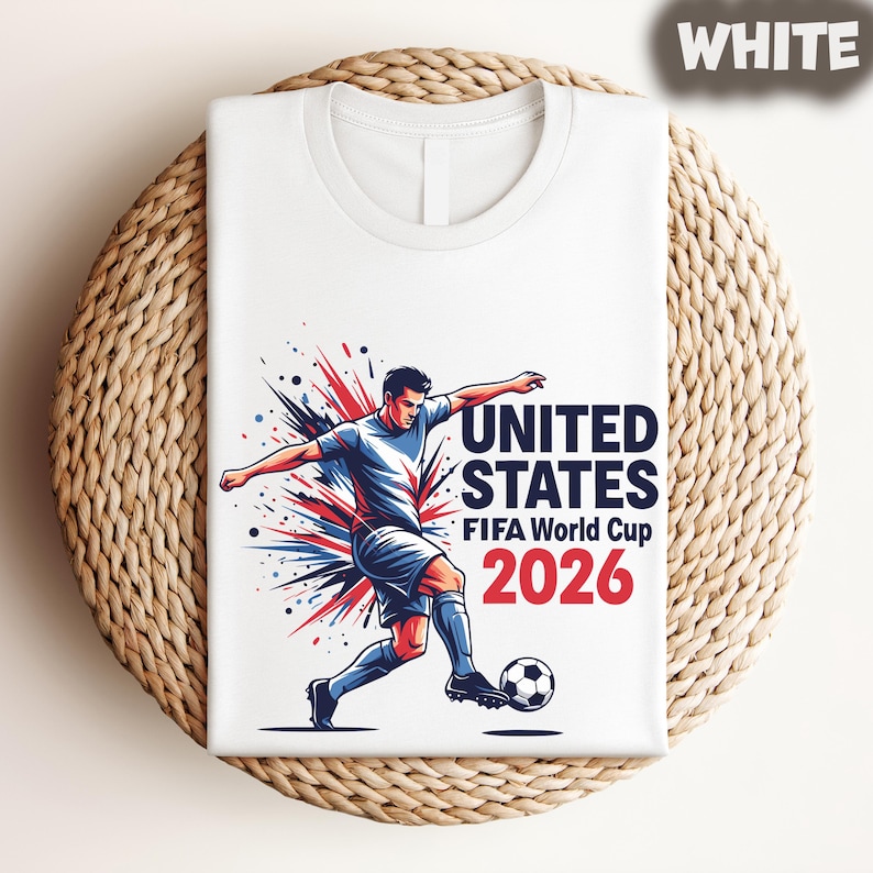 Pode incluir: Camiseta branca com um jogador de futebol a chutar uma bola, com o texto "UNITED STATES FIFA World Cup 2026" em azul marinho e vermelho. Design din&acirc;mico e colorido, ideal para f&atilde;s de futebol.