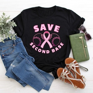 May include: Black t-shirt with the text "SAVE SECOND BASE" in pink lettering. A pink ribbon is centered between two baseballs.