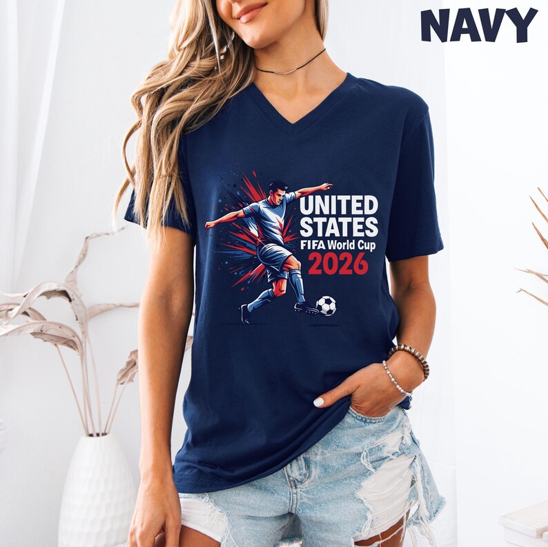 Pode incluir: Camiseta azul marinho com decote em V com um gr&aacute;fico de jogador de futebol e o texto "UNITED STATES FIFA World Cup 2026". O gr&aacute;fico &eacute; em branco e vermelho. A camiseta &eacute; de manga curta.