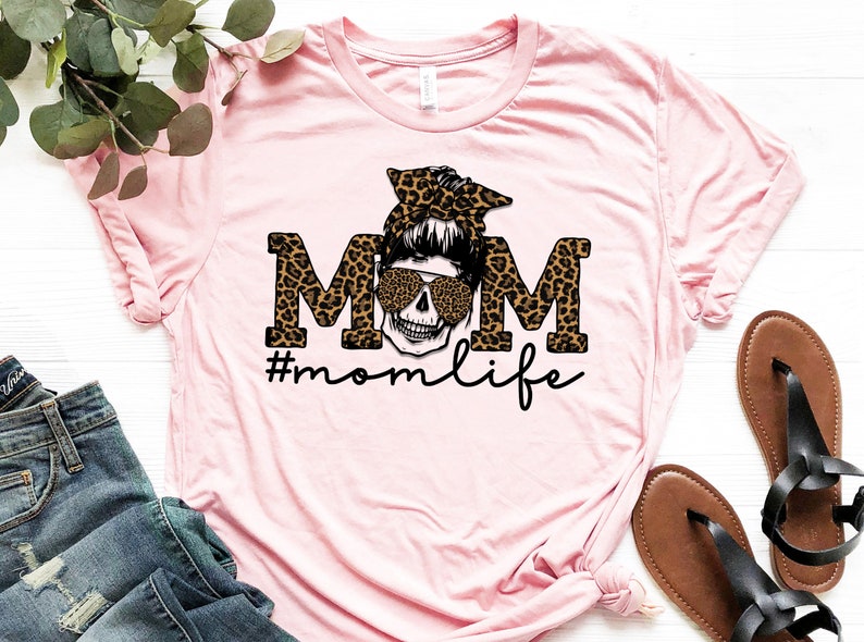May include: Pink t-shirt featuring a black and brown leopard print "MOM" graphic with a skull wearing sunglasses and a headband. The text "#momlife" is below the graphic.