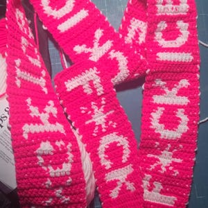 以下が含まれることがあります： 手作りのホットピンクのかぎ針編みのスカーフで、白い文字で「F*CK ICE」と書かれています。スカーフは、テキストと雪の結晶のようなデザインの繰り返しパターンが特徴です。このアイテムは、青いカッティングマットに表示されています。