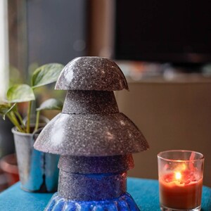 May include: A stack of gray speckled ceramic bowls with a blue light shining from the bottom. The bowls are arranged in a pyramid shape and are sitting on a blue surface. A lit candle is in the background.