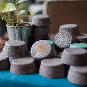 May include: A pile of gray and green round soap bars with a swirl design. The soap bars are stacked on top of each other and are arranged in a pyramid shape.