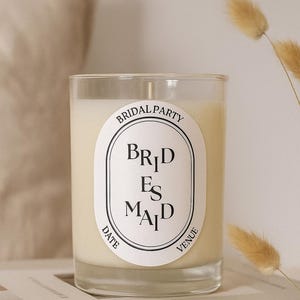 May include: A clear glass candle filled with cream-coloured wax. The candle features a white label with the words "Bridal Party" at the top and "Bridesmaid" in the centre. The words "Date" and "Venue" are at the bottom. The candle rests on a stack of books.