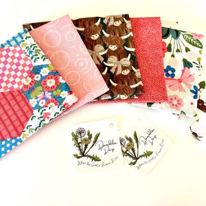 May include: A set of six fabric squares featuring various patterns, including pink and white polka dots, pink and white checkered, floral, and a brown and white highland cow pattern. The fabric squares are arranged on a white surface with two white tags that read "Dagglish Days" and "Where the Granite Green Rises".