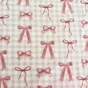 May include: A fabric with a repeating pattern of pink bows on a white and pink gingham background. The bows vary in size and style, some with long flowing ribbons, others tied in neat knots. The overall design is delicate and feminine.