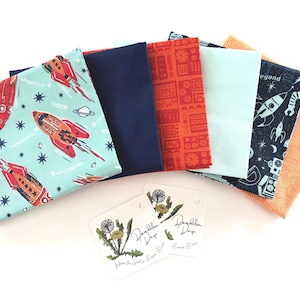 May include: A collection of colorful fabric squares with space-themed patterns, including rockets, planets, and stars. The fabrics are in shades of blue, orange, and teal. Two tags with a dandelion illustration are in the foreground.