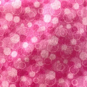 May include: A close-up of a pink fabric with a pattern of white and pink circles. The circles vary in size and are slightly blurred, creating a soft, textured appearance. The background is a solid, vibrant pink.