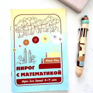 May include: A softcover book with a light blue and yellow cover featuring line drawings of mice, colored circles, and a cake. The text reads "Женя Кац", "ПИРОГ С МАТЕМАТИКОЙ", and "Игры для детей 4-7 лет". Beside it is a multi-colored retractable ballpoint pen.