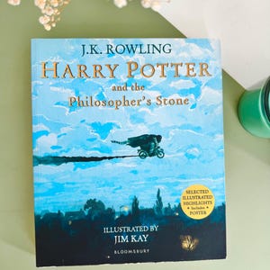 May include: A hardcover book titled Harry Potter and the Philosopher's Stone by J.K. Rowling, illustrated by Jim Kay. The cover features a blue sky with clouds, a figure on a motorcycle, and a yellow circular sticker stating Selected Illustrated Highlights Includes Poster.
