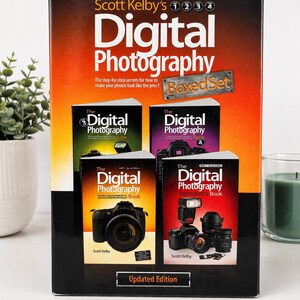 May include: A boxed set of Scott Kelby's Digital Photography books, parts 1 through 4. The black box features an orange gradient background, white text, and images of the four individual books, each displaying a camera and photography equipment.