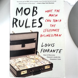 May include: A paperback book titled Mob Rules: What the Mafia Can Teach the Legitimate Businessman by Louis Ferrante. The white cover features a black briefcase filled with stacks of US dollar bills, with red and black text throughout the design.