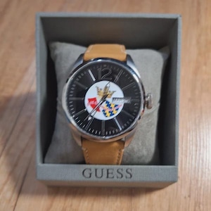May include: A Guess watch with a black face, silver bezel, and tan leather strap. The watch face features a colourful emblem and the number 12. The watch is presented in a grey box.