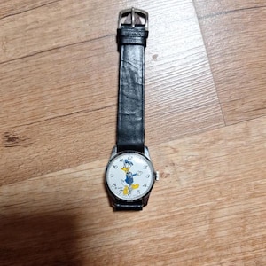 以下が含まれることがあります： 黒い革バンドとシルバーカラーのケースの腕時計。文字盤には、青、黄、白のドナルドダックの漫画の画像が描かれています。バックル留めのクラシックなデザインの時計です。