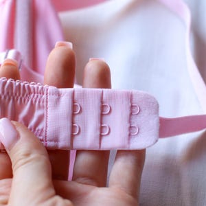 A close-up of a pink bra clasp. The clasp has three sets of hooks and eyes for adjustment. The bra is made of a soft, pink fabric. The clasp is held by a hand with pink nail polish.
