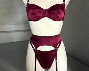 May include: A deep burgundy satin lingerie set featuring a balconette bra with thin adjustable straps, a high-waisted garter belt with scalloped edges, and matching briefs. The set includes attached garter straps with metal hardware and leg loops.