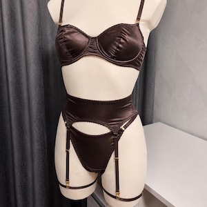 May include: A dark brown satin lingerie set featuring a structured underwire bra with thin adjustable straps, a high-waisted garter belt with a cutout design, and matching briefs. The set includes attached garter straps with gold-toned hardware.